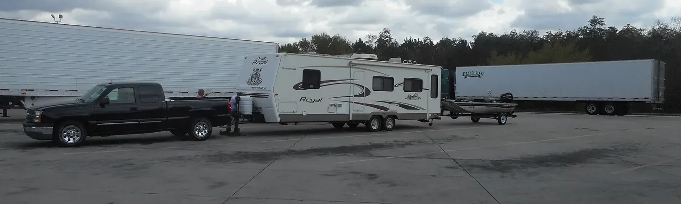 A Chevrolet pickup with a travel trailer and a boat in tow at a highway rest area. A double tow like this is a different setup from the single-trailer examples below.
